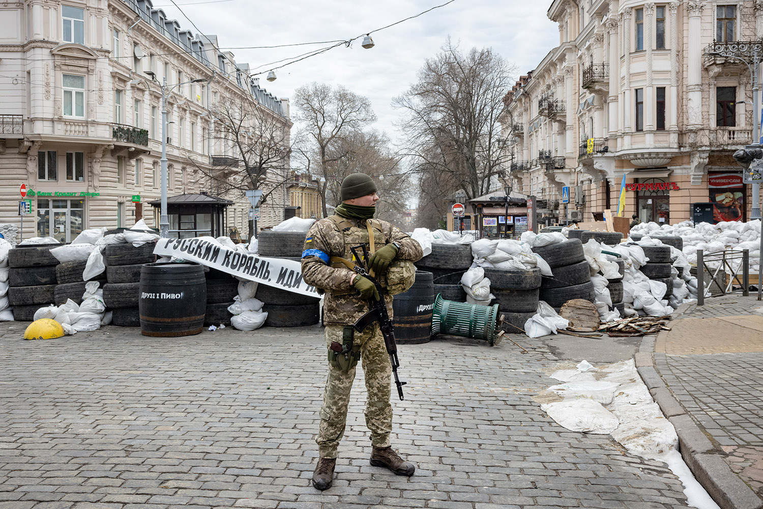 Ukrainian solider standing on a street lined with sandbags, tires, and barrels.